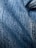 Detailed close-up of blue denim fabric showcasing the texture and weave pattern. The fabric appears slightly wrinkled with visible stitching along one side.