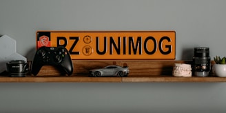 A wooden shelf displays a collection of diverse items including a black game controller, a miniature car model, a yellow and black license plate with the text 'PZ UNIMOG', camera lenses, a model of human teeth, a small potted plant, and a computer networking device.