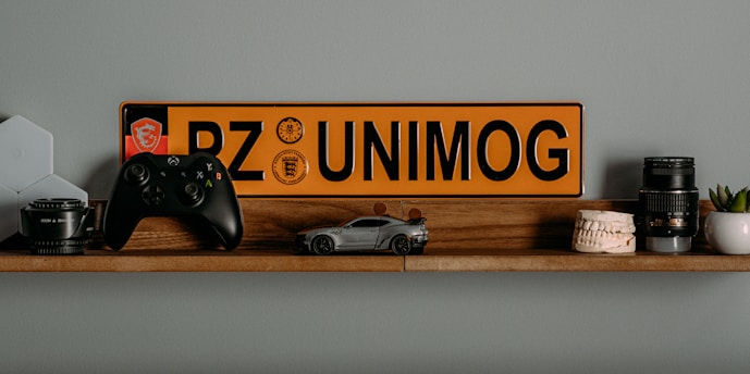 A wooden shelf displays a collection of diverse items including a black game controller, a miniature car model, a yellow and black license plate with the text 'PZ UNIMOG', camera lenses, a model of human teeth, a small potted plant, and a computer networking device.
