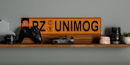A wooden shelf displays a collection of diverse items including a black game controller, a miniature car model, a yellow and black license plate with the text 'PZ UNIMOG', camera lenses, a model of human teeth, a small potted plant, and a computer networking device.