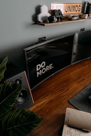 A desktop setup with a motivational message displayed on a wide computer monitor. The monitor is placed on a wooden desk alongside a speaker and a notebook containing handwritten notes. Above the desk, a shelf holds various items including a toy car, a gaming controller, and a series of hexagonal objects. Potted plant leaves slightly encroach into the frame from the left.