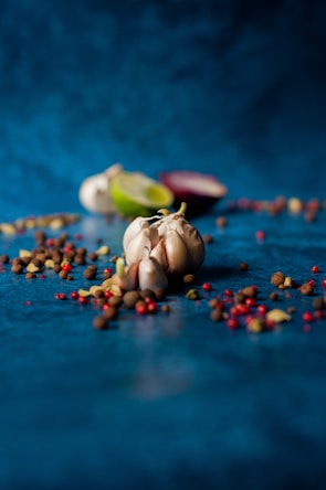 A close-up view of various spices and ingredients scattered on a blue surface. In the foreground, a bulb of garlic sits among assorted whole spices, including black and red peppercorns. A partially cut lime and a halved red onion are blurred in the background, contributing to the culinary theme.