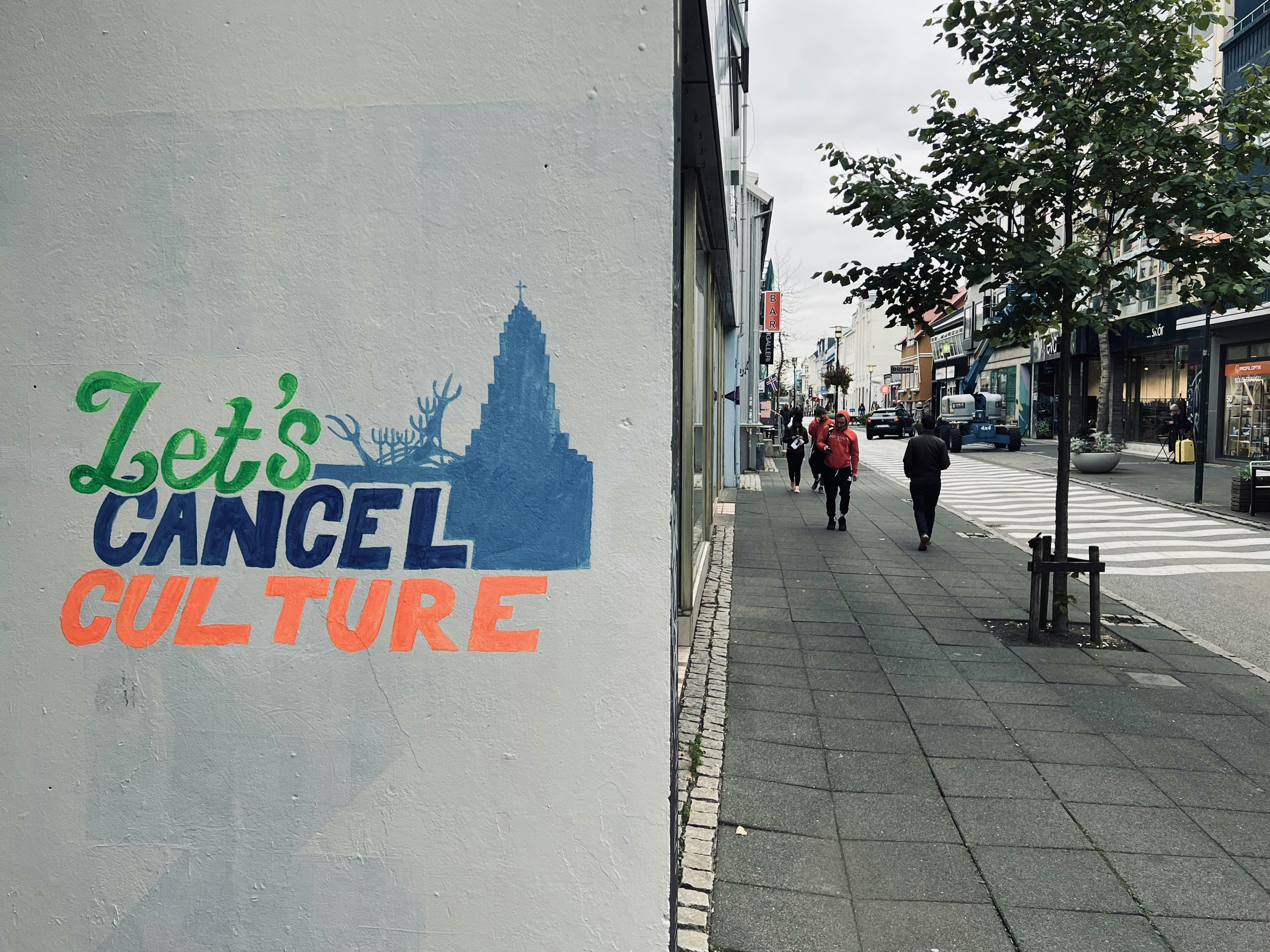 Street scene with a graffiti that reads 'Let's Cancel Culture' in various colors. The graffiti is on a wall beside a paved sidewalk. The street is lined with shops and trees, with pedestrians walking in the distance and cars parked along the road.