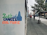 Street scene with a graffiti that reads 'Let's Cancel Culture' in various colors. The graffiti is on a wall beside a paved sidewalk. The street is lined with shops and trees, with pedestrians walking in the distance and cars parked along the road.