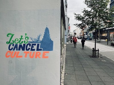 Street scene with a graffiti that reads 'Let's Cancel Culture' in various colors. The graffiti is on a wall beside a paved sidewalk. The street is lined with shops and trees, with pedestrians walking in the distance and cars parked along the road.
