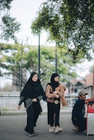 Two women wearing hijabs and casual clothing walk along a pathway lined with trees. A man is seated on the right side, engaging with something out of the frame. The background includes trees and a partially visible structure.