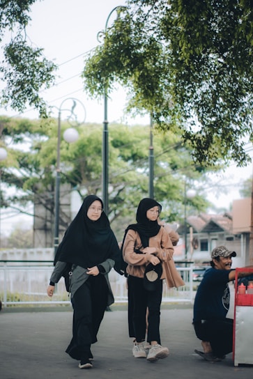 Two women wearing hijabs and casual clothing walk along a pathway lined with trees. A man is seated on the right side, engaging with something out of the frame. The background includes trees and a partially visible structure.