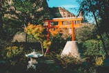 A tranquil garden scene featuring a traditional Japanese torii gate painted in bright red. It stands prominently among lush green foliage and a variety of plants. In the foreground, a stone lantern is surrounded by shrubbery. In the background, rocky cliffs are visible under a clear blue sky.