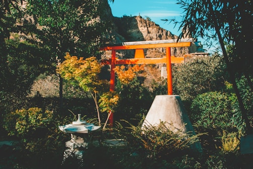 A tranquil garden scene featuring a traditional Japanese torii gate painted in bright red. It stands prominently among lush green foliage and a variety of plants. In the foreground, a stone lantern is surrounded by shrubbery. In the background, rocky cliffs are visible under a clear blue sky.