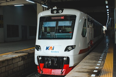 A modern white train with a red base is stationed at a platform, featuring bright lights above. The platform has a yellow safety line and tactile paving. The train is labeled with 'KAI Bandara'.
