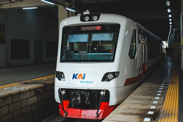 A modern white train with a red base is stationed at a platform, featuring bright lights above. The platform has a yellow safety line and tactile paving. The train is labeled with 'KAI Bandara'.