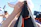 A person wearing a white cap holds a dark piece of clothing with orange stitching, partially covering their face. In the blurry background, another person in a blue swimsuit relaxes in the sun, with beach umbrellas and a blue sky overhead.