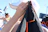 A person wearing a white cap holds a dark piece of clothing with orange stitching, partially covering their face. In the blurry background, another person in a blue swimsuit relaxes in the sun, with beach umbrellas and a blue sky overhead.