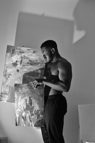 A shirtless man is holding two abstract paintings in a dimly lit room. One painting has a monochromatic palette with abstract brush strokes, and the other depicts hands reaching upwards in a stylized manner. His expression is contemplative, and there is a contrasting play of light and shadow on the wall behind him.