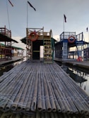 A large bamboo raft with a small structure featuring a ladder and a lifesaver in the center. There are colorful flags attached to tall poles and similar structures on either side. The reflection of the setup is visible in the water beneath, indicating a serene and calm setting, possibly near a body of water like a lake or bay.