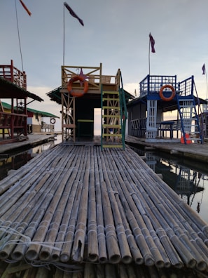 A large bamboo raft with a small structure featuring a ladder and a lifesaver in the center. There are colorful flags attached to tall poles and similar structures on either side. The reflection of the setup is visible in the water beneath, indicating a serene and calm setting, possibly near a body of water like a lake or bay.