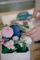A small arrangement of flowers, including pink, purple, and white blossoms, is placed in a rectangular container. A pair of blue scissors lies in the background, and a hand appears to be adjusting or arranging the flowers.