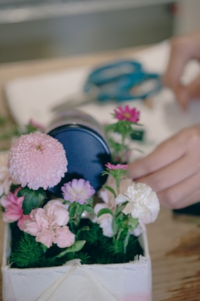 A small arrangement of flowers, including pink, purple, and white blossoms, is placed in a rectangular container. A pair of blue scissors lies in the background, and a hand appears to be adjusting or arranging the flowers.