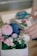 A small arrangement of flowers, including pink, purple, and white blossoms, is placed in a rectangular container. A pair of blue scissors lies in the background, and a hand appears to be adjusting or arranging the flowers.