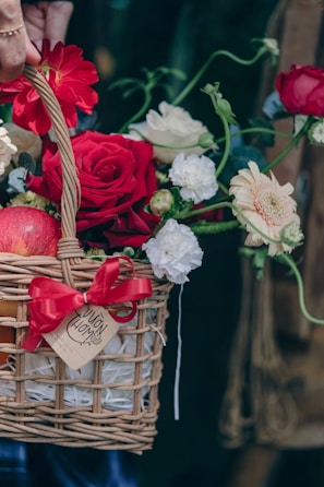 A wicker basket filled with a vibrant arrangement of flowers, including red roses, white carnations, and a red gerbera daisy. A red ribbon is tied around the basket, and there is a tag attached to it. An apple is partially visible inside the basket.