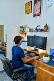 A person is seated at a wooden desk working on a computer with a large monitor displaying code. The setup includes a keyboard, mouse, and a secondary laptop. The walls display motivational posters and a clock. Shelves hold books, headphones, and a Buddha sculpture. There is a potted plant on the desk. The environment appears organized and conducive to focused work.