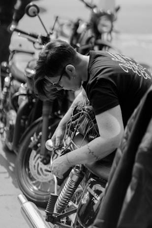 A person with short hair is working on or inspecting a motorcycle, surrounded by multiple bikes. They seem focused, with tools or gear possibly in their hands. The image is in black and white, highlighting the contrast and details in the machinery.