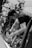 A person with short hair is working on or inspecting a motorcycle, surrounded by multiple bikes. They seem focused, with tools or gear possibly in their hands. The image is in black and white, highlighting the contrast and details in the machinery.