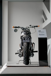 A vintage motorcycle is displayed indoors, positioned on a platform. The motorcycle is viewed from the rear, showcasing a black license plate that reads 'FRX 4048'. The background is minimalistic, with geometric patterns on the wall to the right.