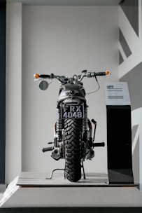 A vintage motorcycle is displayed indoors, positioned on a platform. The motorcycle is viewed from the rear, showcasing a black license plate that reads 'FRX 4048'. The background is minimalistic, with geometric patterns on the wall to the right.
