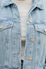 A close-up of a light blue denim jacket featuring metallic buttons. The jacket is layered over a white ribbed top, partially visible.