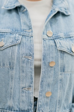 A close-up of a light blue denim jacket featuring metallic buttons. The jacket is layered over a white ribbed top, partially visible.