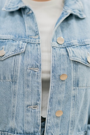 A close-up of a light blue denim jacket featuring metallic buttons. The jacket is layered over a white ribbed top, partially visible.