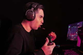 A person wearing headphones is intensely focused on a computer screen while playing a video game with a controller in hand. The dimly lit environment suggests a gaming setup with a visible screen displaying a game.