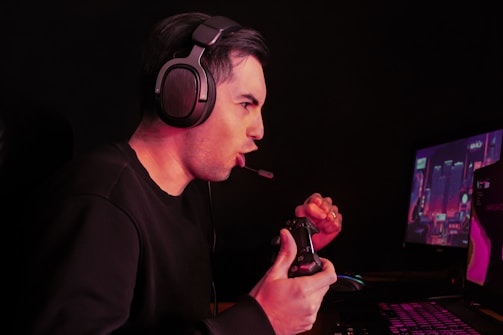 A person wearing headphones is intensely focused on a computer screen while playing a video game with a controller in hand. The dimly lit environment suggests a gaming setup with a visible screen displaying a game.