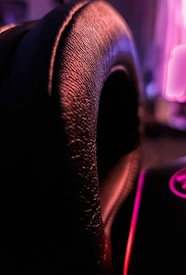 Close-up of a textured black leather surface with dim, moody lighting creating a dramatic effect. The background is blurred, but hints of pink and purple neon-like lights are visible.