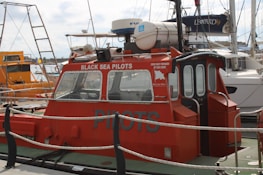 A red pilot boat is docked at a marina, with 'Black Sea Pilots' written on the side. The boat features windows at the front and a small enclosed cabin. Various ropes and a safety rail are visible around the vessel. Other boats and sailing masts are in the background, including a larger, white boat labeled 'Leopard 39'. The sky is cloudy.