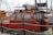 A red pilot boat is docked at a marina, with 'Black Sea Pilots' written on the side. The boat features windows at the front and a small enclosed cabin. Various ropes and a safety rail are visible around the vessel. Other boats and sailing masts are in the background, including a larger, white boat labeled 'Leopard 39'. The sky is cloudy.