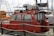 A red pilot boat is docked at a marina, with 'Black Sea Pilots' written on the side. The boat features windows at the front and a small enclosed cabin. Various ropes and a safety rail are visible around the vessel. Other boats and sailing masts are in the background, including a larger, white boat labeled 'Leopard 39'. The sky is cloudy.