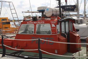 A red pilot boat is docked at a marina, with 'Black Sea Pilots' written on the side. The boat features windows at the front and a small enclosed cabin. Various ropes and a safety rail are visible around the vessel. Other boats and sailing masts are in the background, including a larger, white boat labeled 'Leopard 39'. The sky is cloudy.