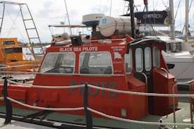 A red pilot boat is docked at a marina, with 'Black Sea Pilots' written on the side. The boat features windows at the front and a small enclosed cabin. Various ropes and a safety rail are visible around the vessel. Other boats and sailing masts are in the background, including a larger, white boat labeled 'Leopard 39'. The sky is cloudy.