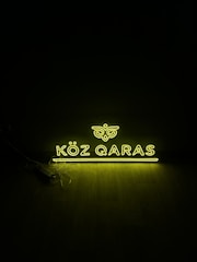 A glowing neon sign displaying the words 'KÖZ QARAS' in bright yellow light against a dark background. The light casts a subtle glow on the floor beneath, and some cables are visible beside the sign.