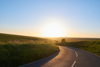 A winding back road cutting through golden fields under a wide blue sky at sunset.