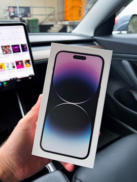 A person holds a smartphone box inside a car. The box features an image of a phone with a gradient screen effect in shades of purple and blue. The interior of the car is modern with a visible tablet-like display on the dashboard, showing various app icons.