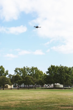 A drone is flying high in the sky above a large, open grassy field. In the background, there is a row of trees with a few buildings partially obscured by the foliage. The sky is mostly clear with some clouds scattered across it.