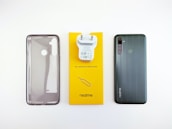 A phone case, phone charger, and a smartphone are placed side by side on a white surface. The phone case is translucent with cutouts for the camera and fingerprint sensor. The charger is white and attached to a bright yellow card that has small text and the brand logo. The smartphone is black with a triple camera setup, fingerprint sensor, and brand logo on the back.