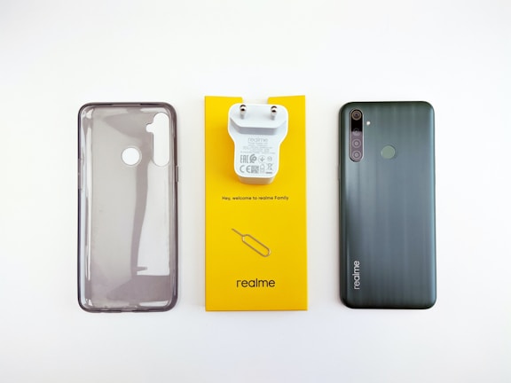 A phone case, phone charger, and a smartphone are placed side by side on a white surface. The phone case is translucent with cutouts for the camera and fingerprint sensor. The charger is white and attached to a bright yellow card that has small text and the brand logo. The smartphone is black with a triple camera setup, fingerprint sensor, and brand logo on the back.