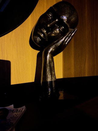 A dark, metallic sculpture of a human face resting on a hand, set against a warm, wooden background. The sculpture's reflective surface catches the light, creating contrasting highlights and shadows. A magazine with partially visible text lies on a dark surface nearby.