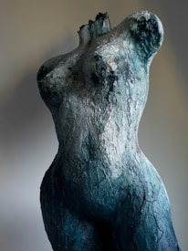 A textured sculpture of a female torso, made to resemble cracked and weathered stone. The sculpture is rough and uneven, with deep grooves and crevices, lending it a rugged appearance.