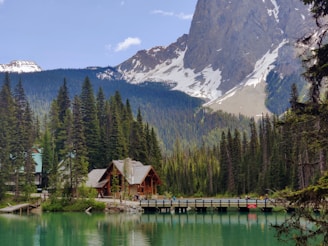 A serene mountain landscape features a wooden cabin surrounded by dense evergreen trees and a lake with turquoise water. A bridge crosses the lake, and snow-capped peaks are visible in the background.