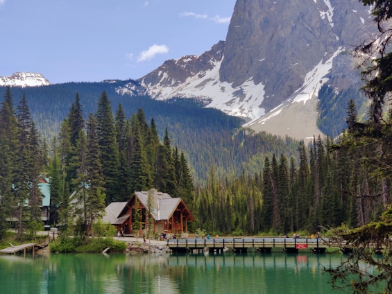 A serene mountain landscape features a wooden cabin surrounded by dense evergreen trees and a lake with turquoise water. A bridge crosses the lake, and snow-capped peaks are visible in the background.